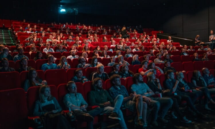 A large group of people seated in red chairs inside a movie theater, watching a film on a screen at the front.