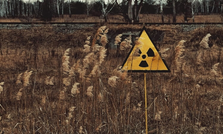 A yellow triangular radiation warning sign stands in tall dry grass in a barren outdoor area with trees and a railway in the background.