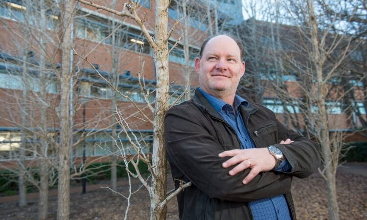 A man in a jacket stands with arms crossed in front of leafless trees and a brick building.