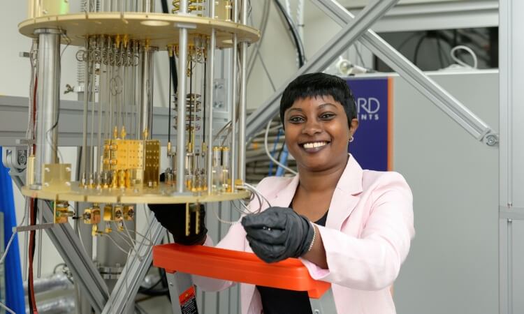 A woman in a light pink blazer and black gloves stands on a ladder, adjusting components of a quantum computing device in a laboratory.