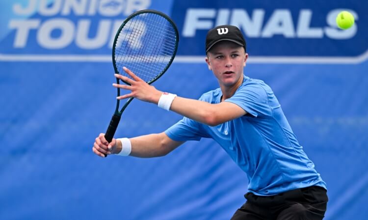 A young tennis player in a blue shirt and black cap prepares to hit a forehand shot during a match, with a "Junior Tour Finals" banner in the background.