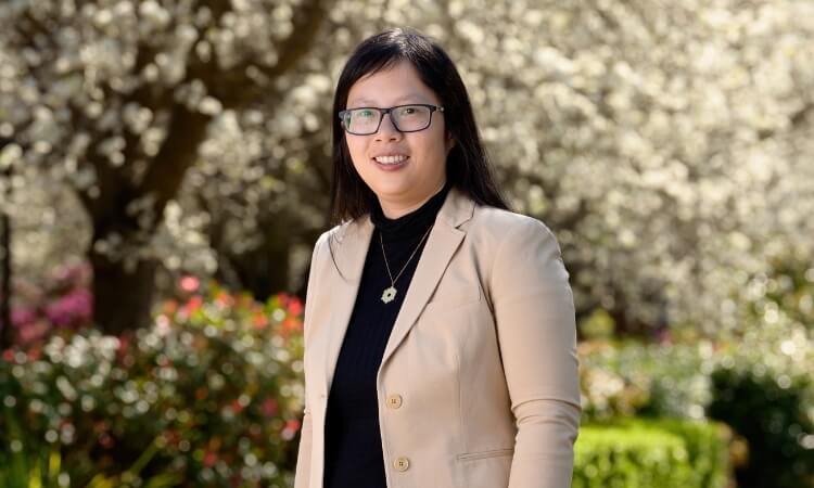A woman with glasses, wearing a beige blazer and black top, stands outdoors in front of blooming trees and greenery.