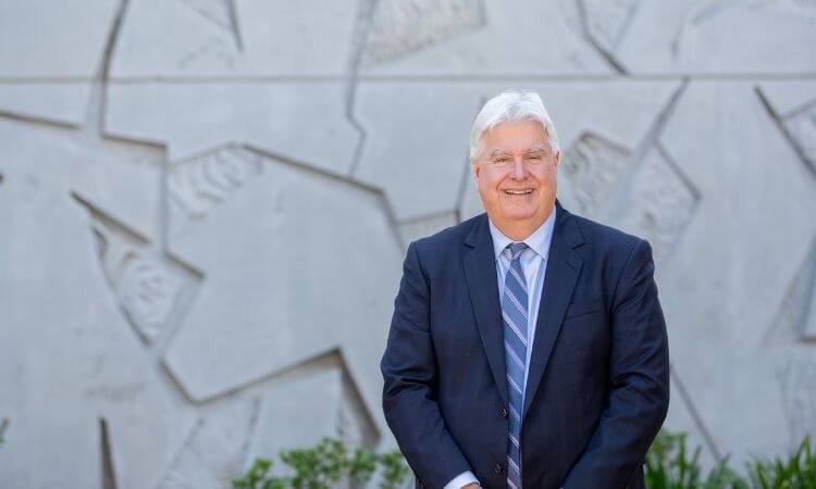 An older man in a dark suit and striped tie stands outside in front of a patterned concrete wall, smiling at the camera.
