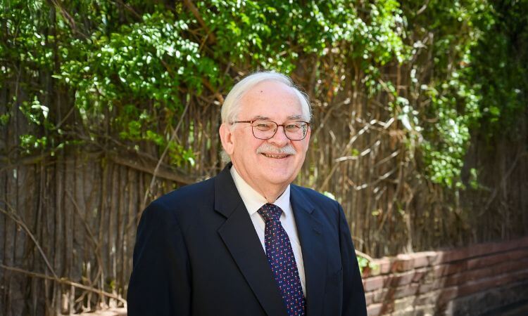An older man in a suit and tie stands outdoors in front of a wall covered with green vines, smiling at the camera.