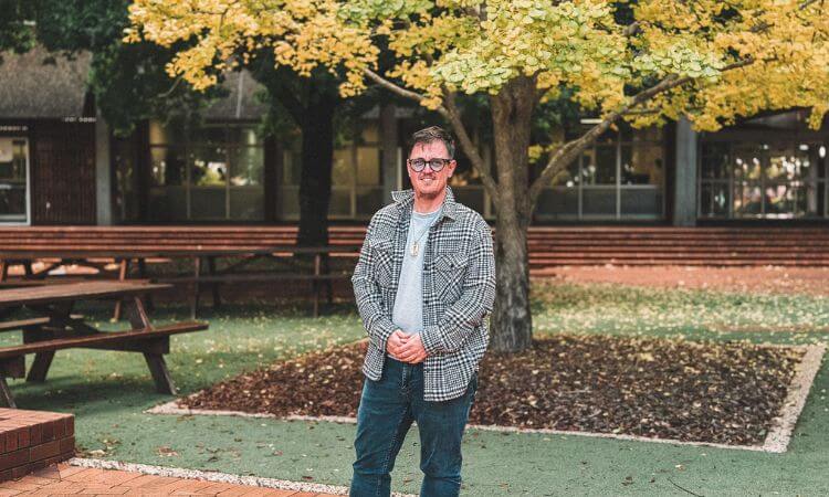 A person wearing glasses and a plaid jacket stands in front of a tree with yellow leaves in an outdoor area with benches.