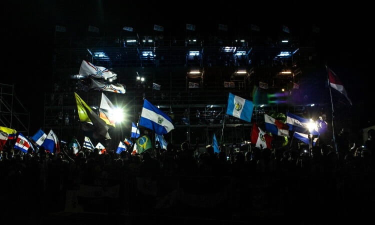 A crowd holds various national flags at night in front of a brightly lit stage structure.