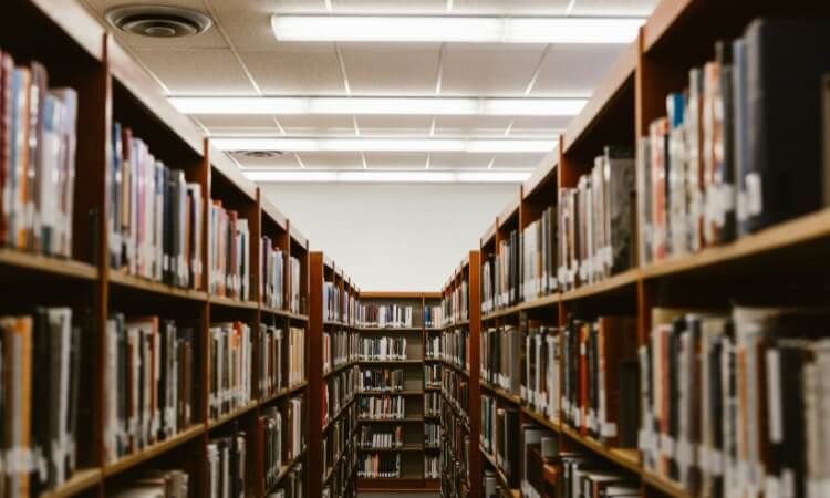 Rows of bookshelves filled with books line a library aisle under fluorescent ceiling lights.