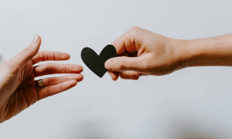 One hand passes a small black paper heart to another outstretched hand against a plain white background.