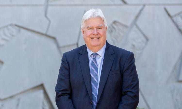 An older man with white hair and glasses wearing a dark suit, blue shirt, and striped tie stands smiling in front of a textured concrete wall.