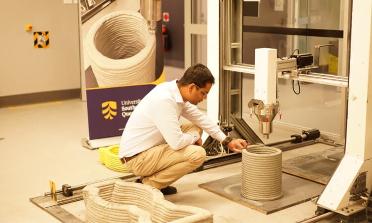 A man examines a cylindrical concrete structure being 3D printed by a large machine in a laboratory setting.