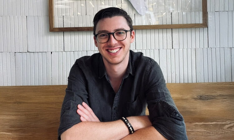 A man wearing glasses and a dark shirt sits at a wooden table, smiling with his arms folded in front of him against a textured white wall.