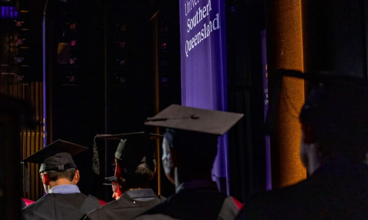 Graduates in caps and gowns stand facing a stage with a University of Southern Queensland banner in the background.