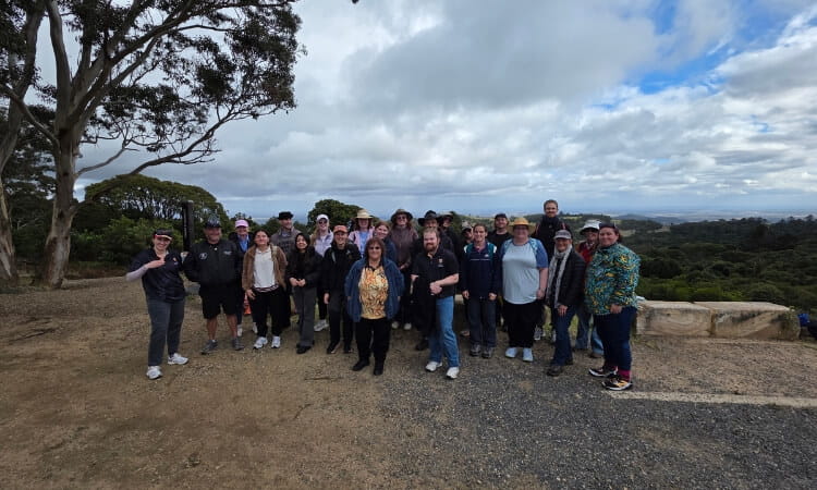 A group of people pose for a photo outdoors on a dirt path, with trees, greenery, and a cloudy sky in the background.