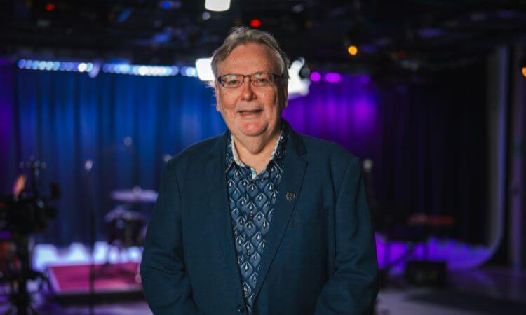 A man wearing glasses and a blue blazer stands smiling in a studio with purple and blue stage lights in the background.