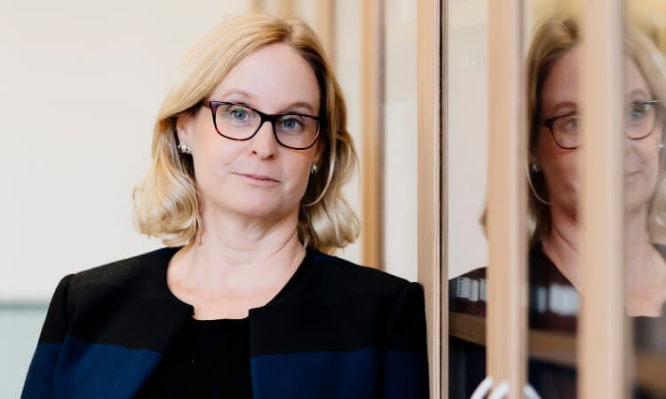 A woman with blonde hair and glasses stands indoors next to a glass wall, her reflection visible beside her. She is wearing a dark jacket and looking at the camera with a neutral expression.