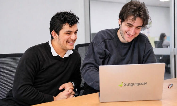 Two men sit at a table, smiling while looking at a laptop with the "Gutgutgoose" logo in an office setting.
