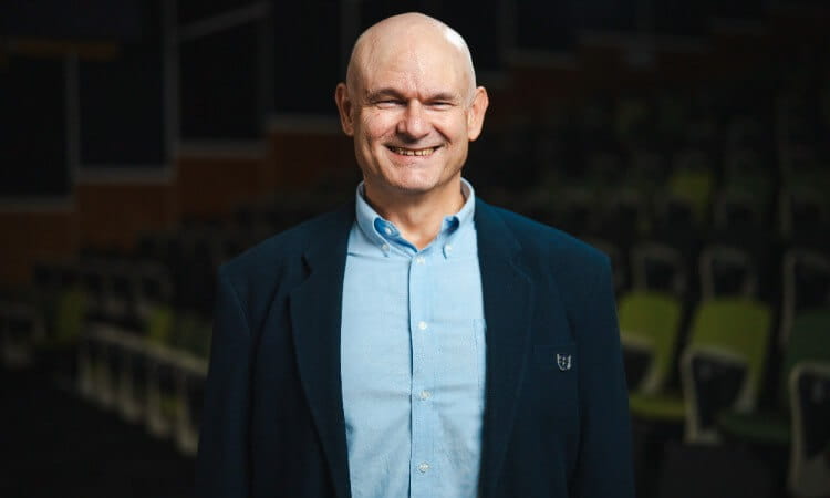 A bald man wearing a blue shirt and dark blazer stands indoors, smiling at the camera with rows of chairs in the background.