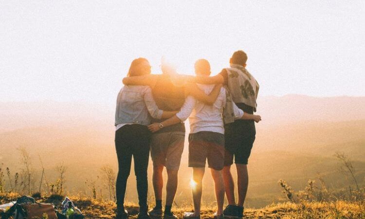 Four people stand side by side with arms around each other, facing a sunset over a landscape of rolling hills.