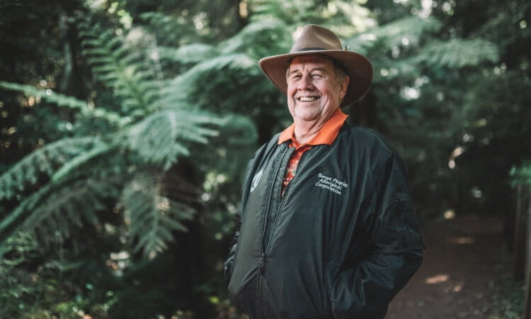 An older man wearing a wide-brimmed hat and a dark jacket smiles while standing on a forest path surrounded by greenery.