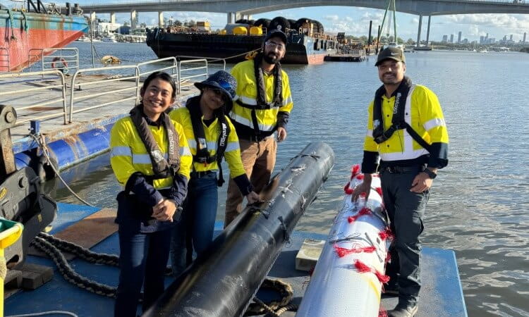 Four people in high-visibility workwear stand on a dock by the water, handling large cylindrical equipment with boats and a bridge in the background.