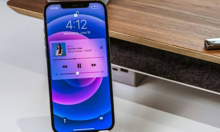 An iPhone displaying the lock screen at 4:12 with a music player showing "favorite crime" by Olivia Rodrigo is placed on a stand near a wooden desk.