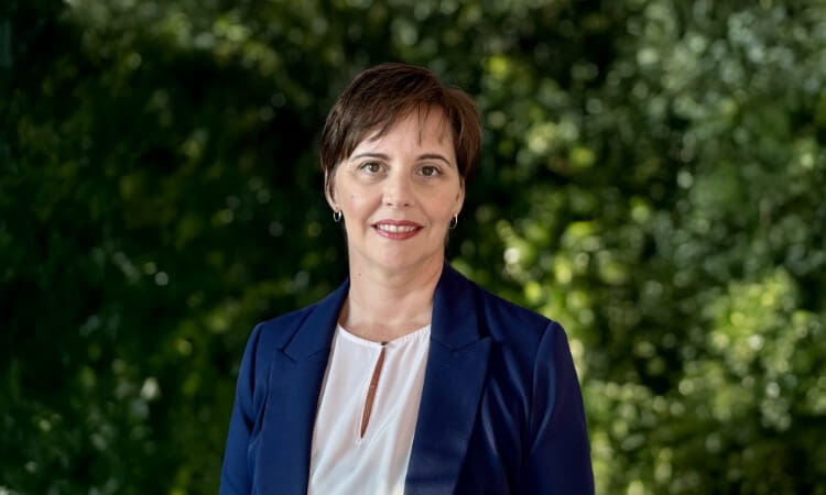A woman with short brown hair wearing a blue blazer and white blouse stands outdoors in front of green foliage, smiling at the camera.