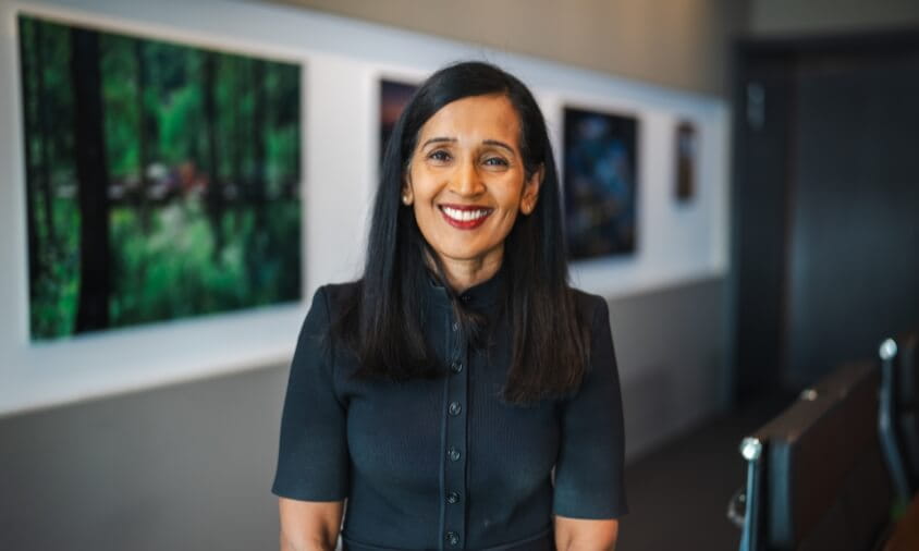 A woman with long dark hair and a black dress smiles while standing in a modern hallway with framed photos on the wall behind her.