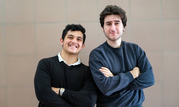 Two young men stand side by side, smiling at the camera with their arms crossed, in front of a plain tiled wall.
