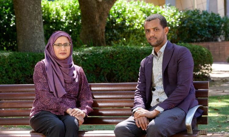 Two people sit on a wooden bench in an outdoor setting with trees and bushes in the background. Both are looking at the camera.