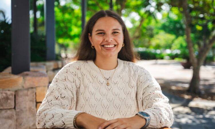 A woman with long brown hair, wearing a cream-colored knit sweater and jewelry, sits at an outdoor table and smiles at the camera with trees in the background.