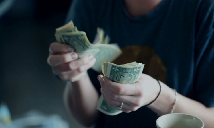 Person wearing a dark shirt counts a stack of one-dollar bills with both hands, with a cup partly visible in the foreground.