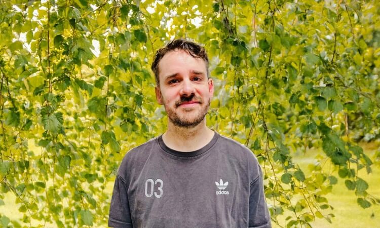 A person in a dark Adidas t-shirt stands outside in front of leafy green branches, looking at the camera.