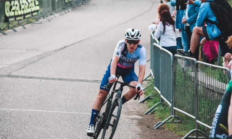 A cyclist wearing a helmet and sunglasses rounds a corner during a race, with spectators standing behind metal barriers along the road.