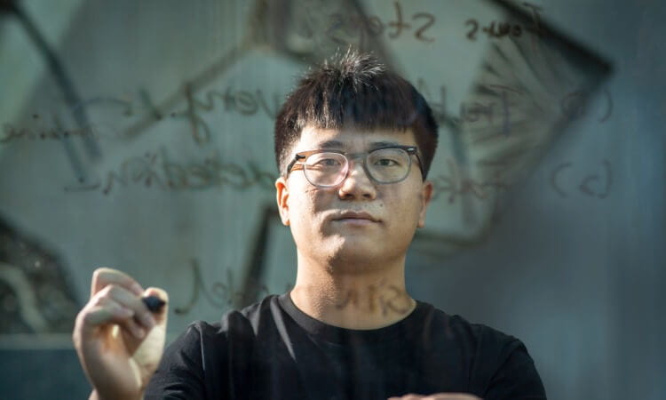 A person wearing glasses writes with a marker on a transparent surface covered in handwritten text, facing the camera.