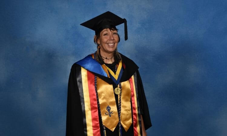 A person wearing a graduation cap and gown with multiple honor cords and sashes stands in front of a blue backdrop, smiling at the camera.