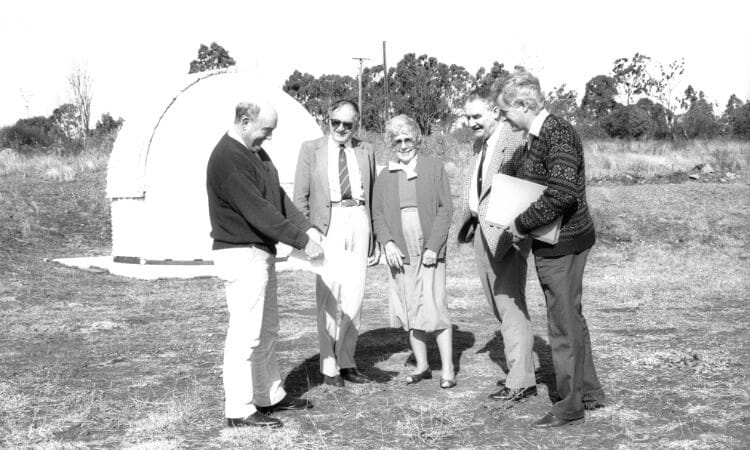 Five people stand on dry grass near a small dome-shaped building, engaged in conversation and holding papers, with trees in the background.