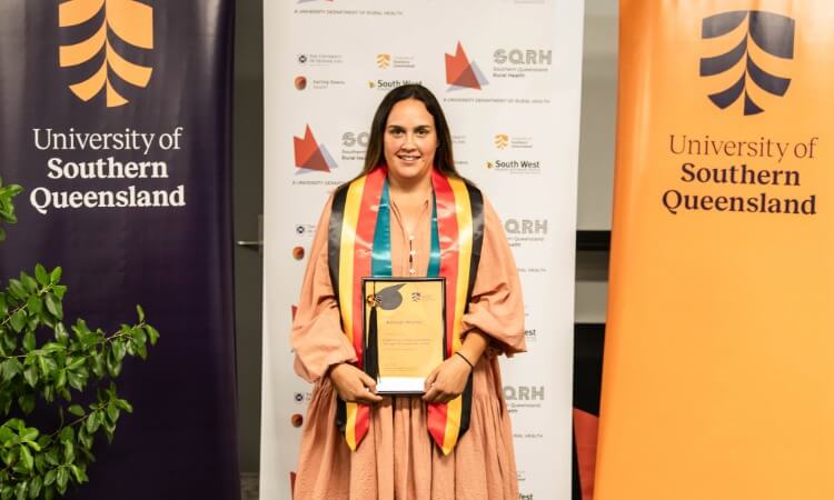 A woman in a pink dress holds a certificate and wears a graduation stole, standing between University of Southern Queensland banners.