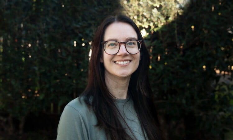 A woman with long dark hair and glasses smiles at the camera while standing outdoors with greenery in the background.