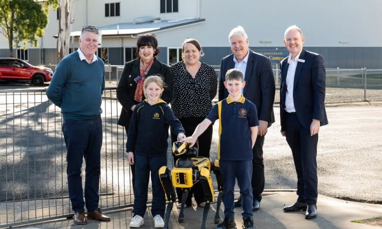 Two children and a yellow robot dog stand in front of four smiling adults in business attire, outdoors near a building and fencing.
