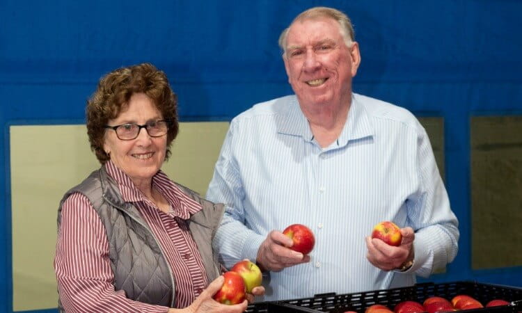 Two older adults stand indoors, smiling and holding apples from a black crate in front of a blue background.