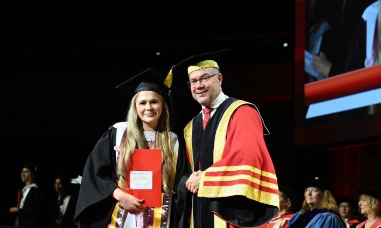 A graduate in cap and gown holds a certificate while standing next to an official in ceremonial academic attire at a graduation ceremony.