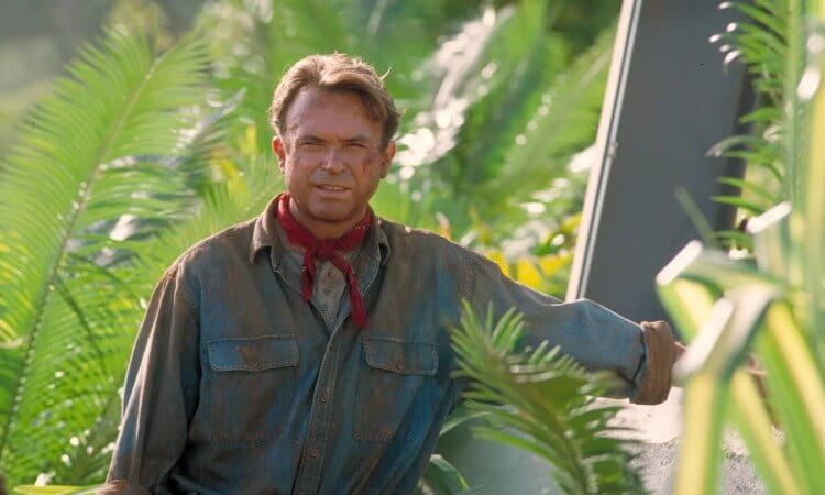 A man in a weathered shirt and red bandana stands among lush green foliage, with dirt on his face and a serious expression, outdoors in a jungle setting.