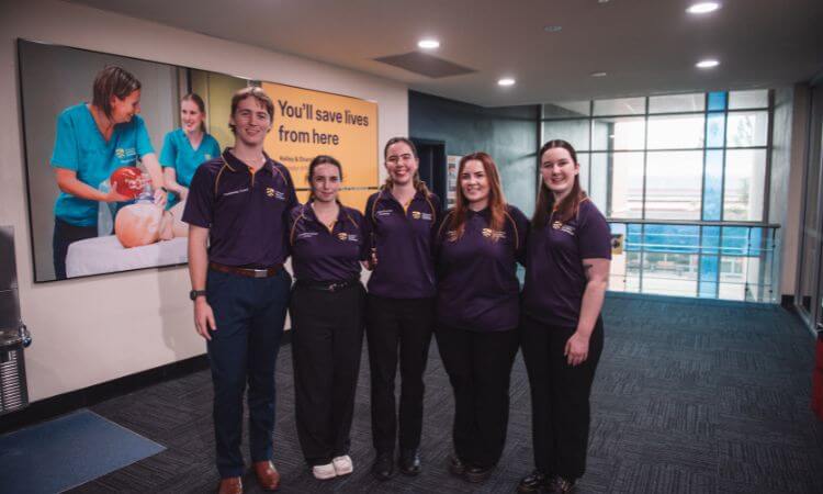 Five people in matching purple shirts stand together in a hallway, with a poster about saving lives visible on the wall behind them.