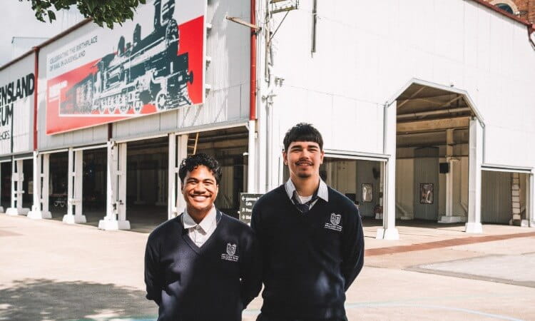 Two students in dark school uniforms stand smiling outside a building with a large train mural and “Queensland Museum” signage visible.