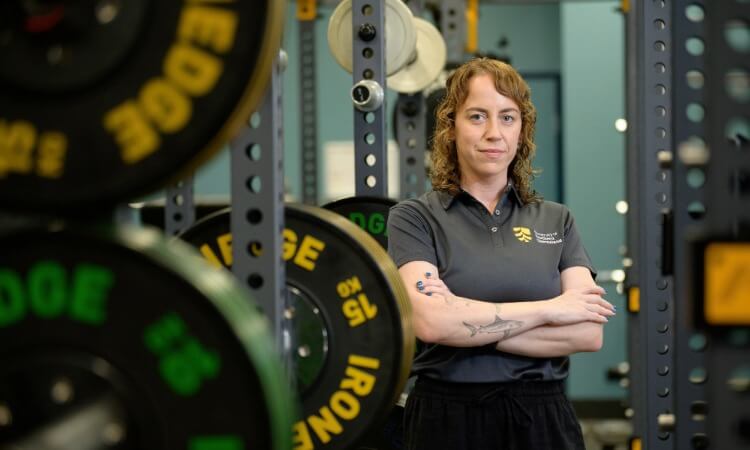 A person with curly hair stands with arms crossed in a gym, surrounded by weightlifting equipment and colorful weight plates.