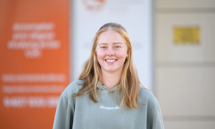 A young woman with long blonde hair, wearing a gray hoodie, smiles at the camera, standing in front of a blurred orange and white background.