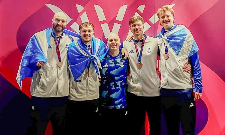 Five athletes stand together, some wearing medals and blue flags over their shoulders, in front of a purple and pink backdrop with a white logo.