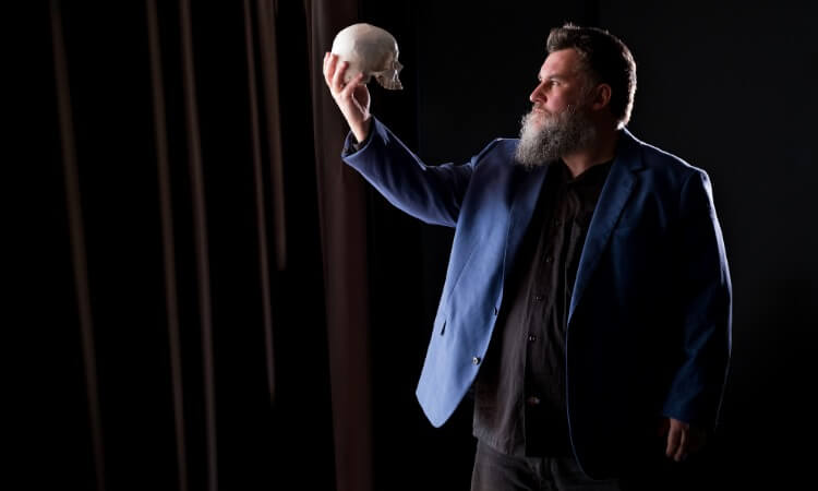 A man with a beard in a blue blazer holds up a human skull and looks at it against a dark background.