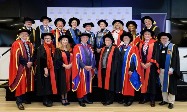 A group of people in academic regalia pose together indoors, wearing graduation gowns, caps, and sashes, standing in front of a logo-covered backdrop.
