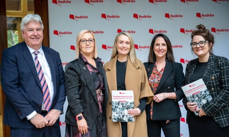 Five people stand in front of a Redkite-branded backdrop, three holding Redkite Financial Crisis booklets, all dressed in business attire, posing for a group photo.